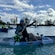 A man sitting in a kayak while holding up a fishing pole and a fish he has caught